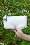 photo shows a white crossbody/ wristlet in front of greenery.