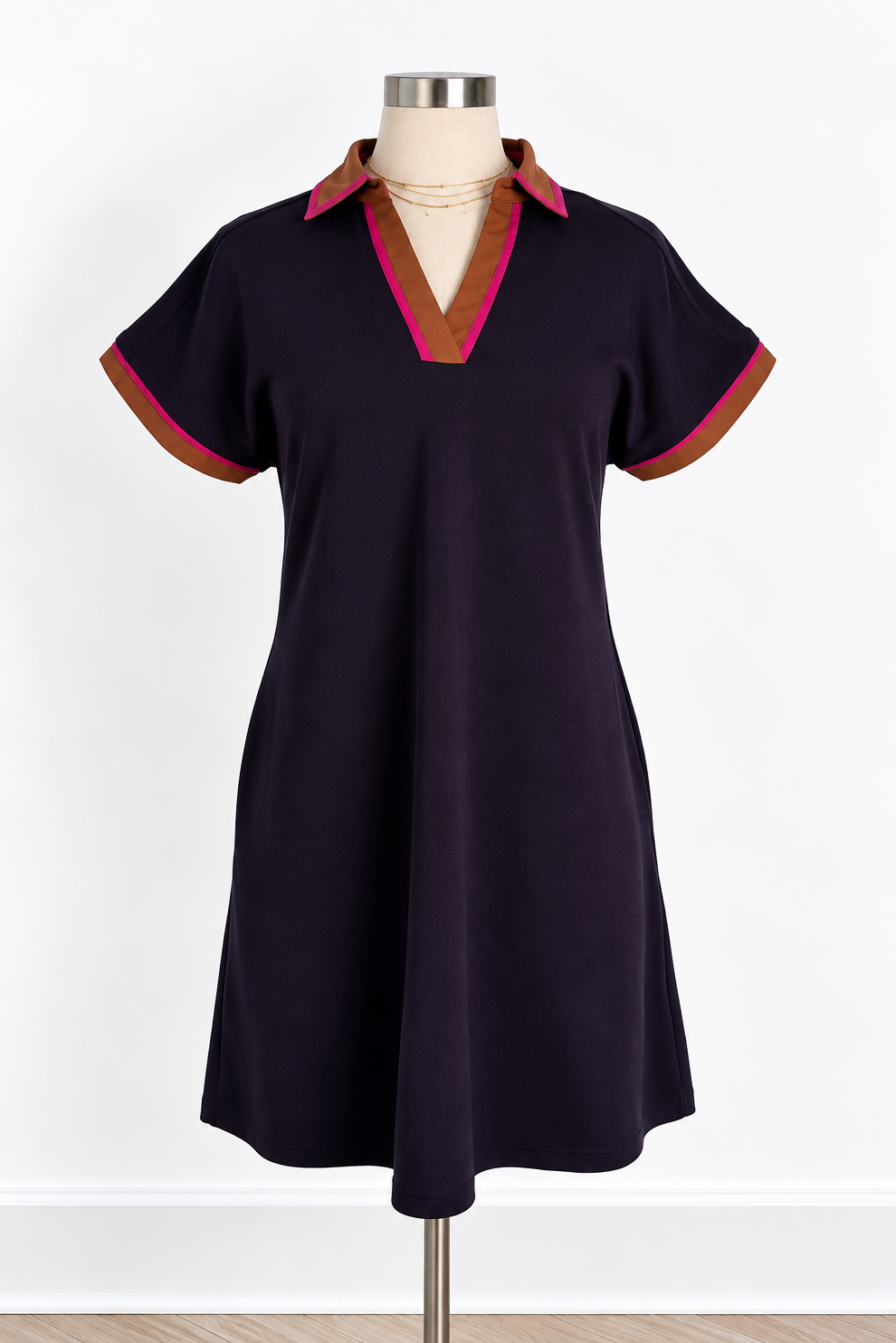 A short-sleeve polo-style dress displayed on a mannequin against a white wall. The dress is dark navy with contrasting brown and pink trim on the collar and sleeve cuffs.
