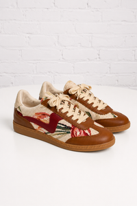 A pair of low-top sneakers with brown leather toe caps and quarter panels, beige knit uppers featuring a red and green floral pattern, beige laces, and brown gum soles, set against a white brick wall background.