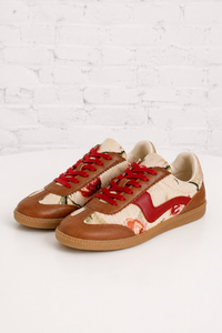 A pair of low-top sneakers with brown leather panels, a cream and orange floral print upper, and bright red laces, displayed on a white surface against a white brick wall background.
