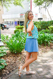 Model is wearing a blue paisley tank with a blue mini skirt and white strappy sandals in front of greenery.