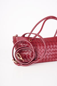 A close-up of a woven pleather handbag in deep burgundy red, featuring a matching coiled shoulder strap with gold-tone hardware, set against a plain white background.