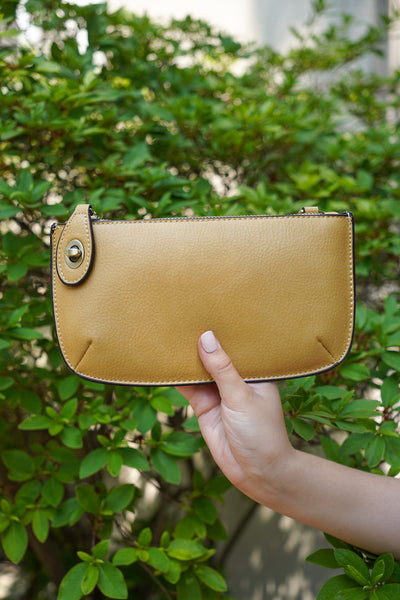 photo shows a mustard color crossbody/ wristlet in front of greenery.