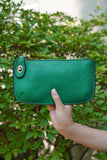 photo shows a green crossbody/ wristlet in front of greenery.