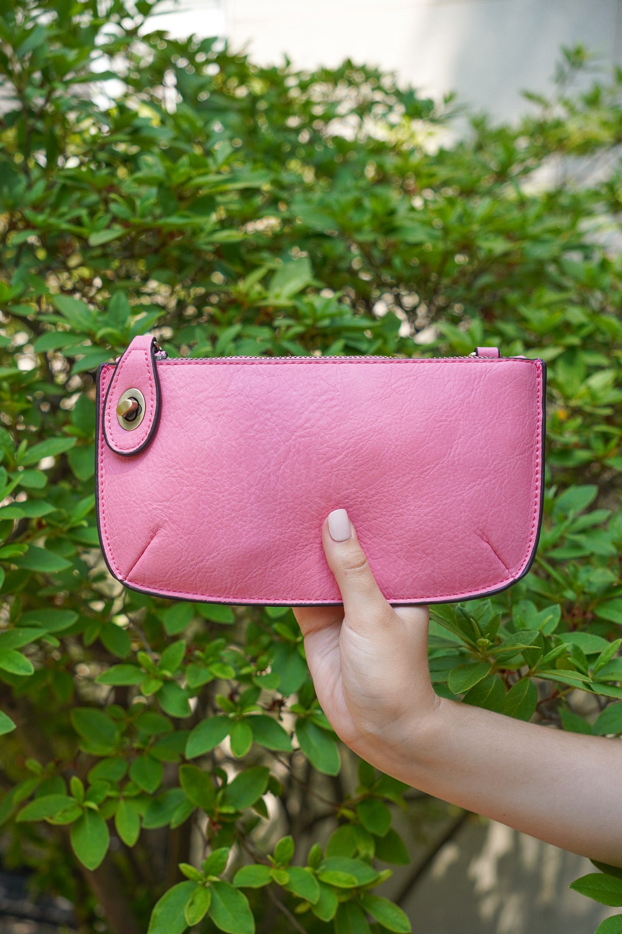 photo shows a pink crossbody/ wristlet in front of greenery.