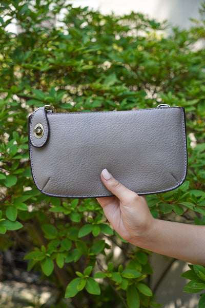photo shows a grey crossbody/ wristlet in front of greenery.
