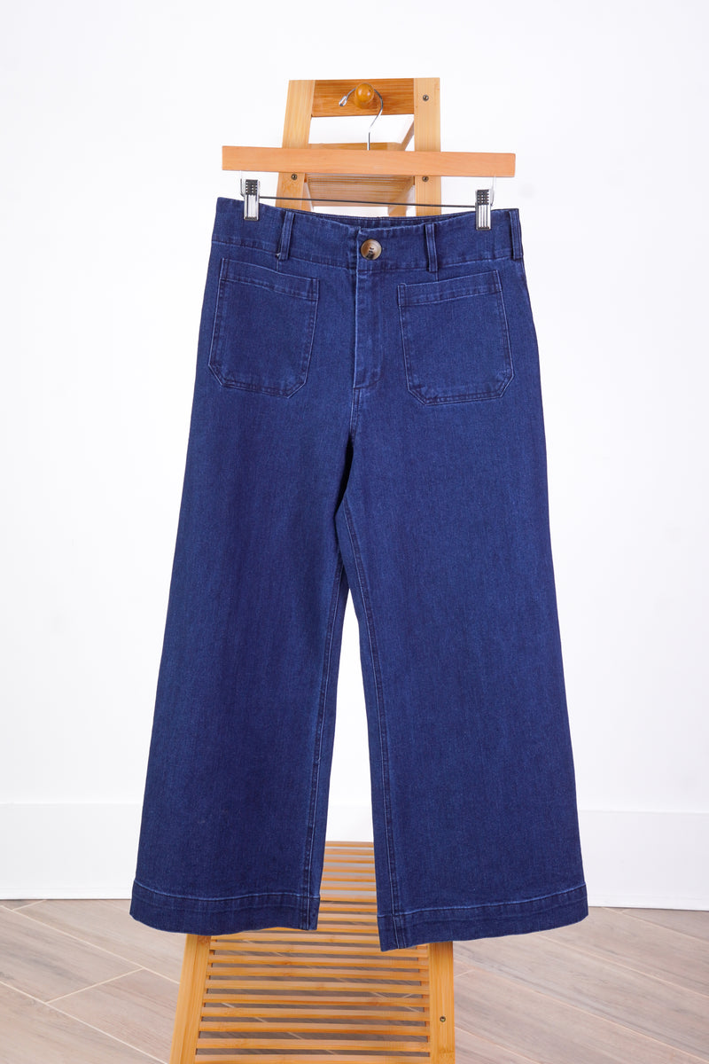 A pair of dark blue wide-leg denim pants hanging from a wooden hanger stand against a white wall and light wood floor.