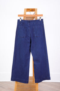 A pair of dark blue wide-leg denim pants shown from the back, hanging from a wooden hanger on a wooden stand against a white wall.