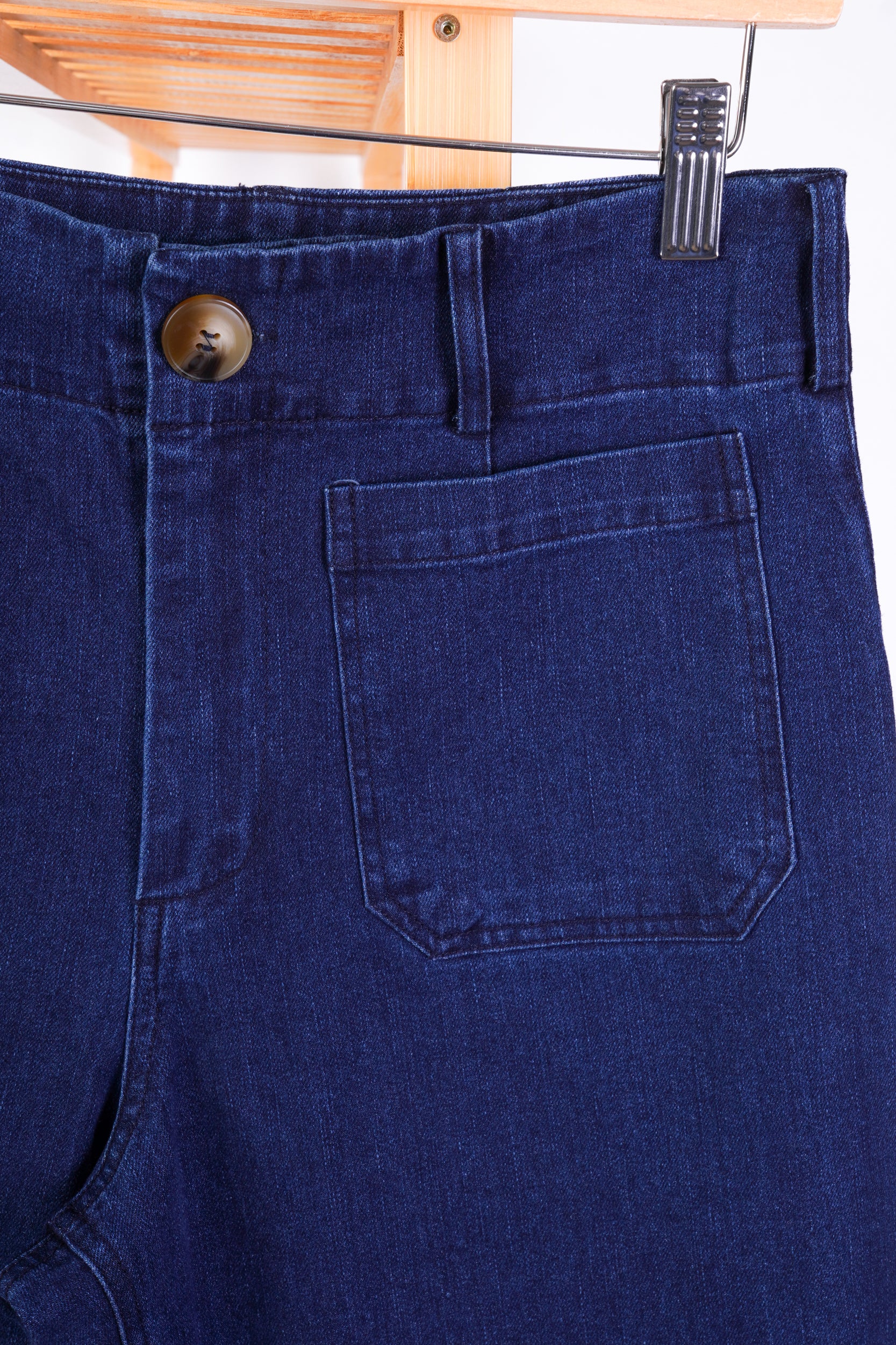 Close-up of dark blue denim jeans showing the waistband, button closure, belt loops, and a front patch pocket, hanging on a wooden hanger.