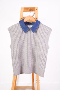 A light gray cable-knit sweater vest with a contrasting blue denim collar, hanging on a wooden hanger attached to a wooden step ladder against a white wall.