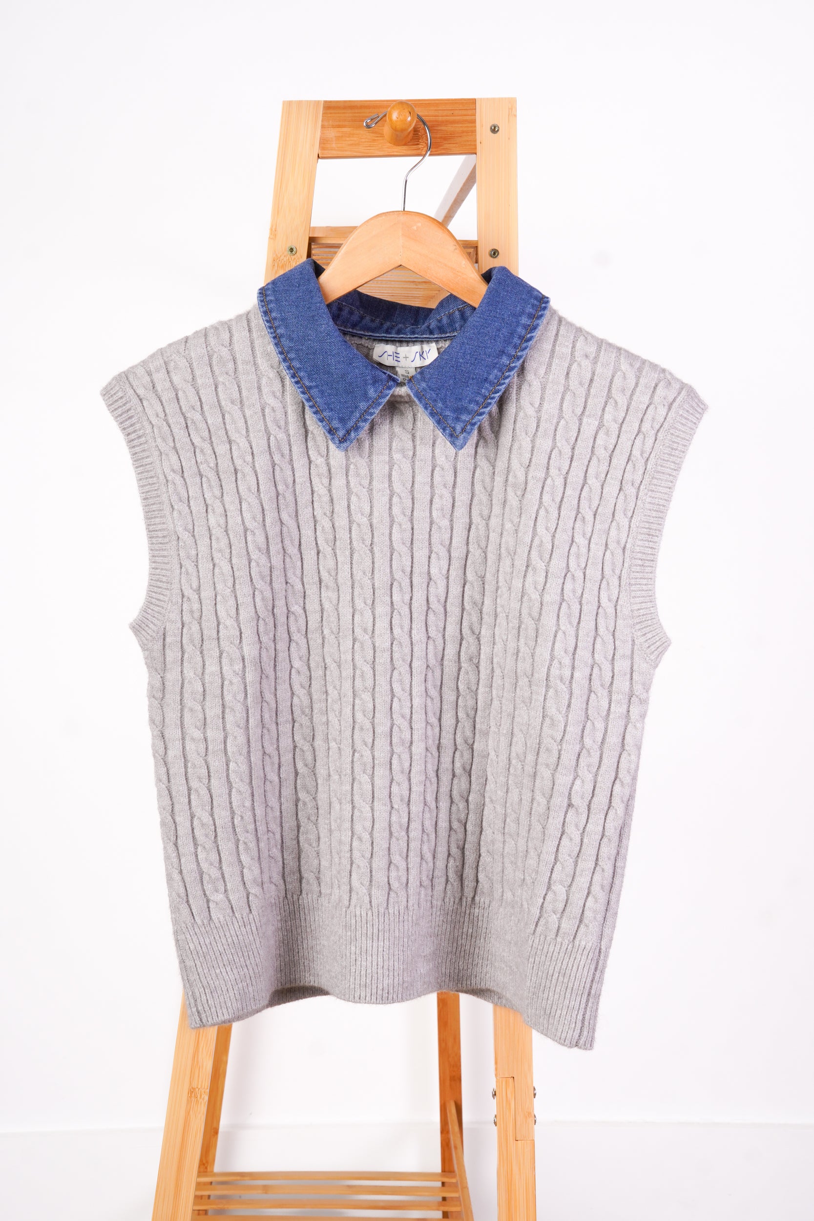 A light gray cable-knit sweater vest with a contrasting blue denim collar, hanging on a wooden hanger attached to a wooden step ladder against a white wall.