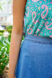 Model is wearing a blue paisley print tank with a blue denim washed skirt in front of greenery.