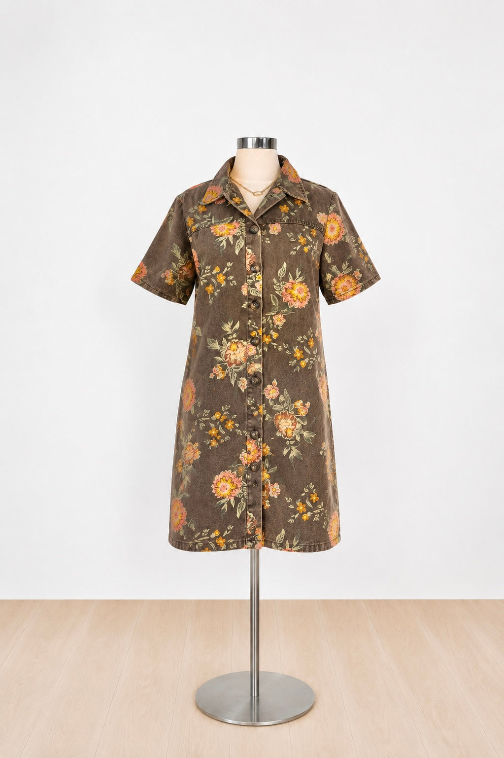 A short-sleeve button-front shirt dress displayed on a mannequin stand. The dress has a brown base with a retro floral pattern in orange, yellow, and green, set against a plain white wall and light wood floor.