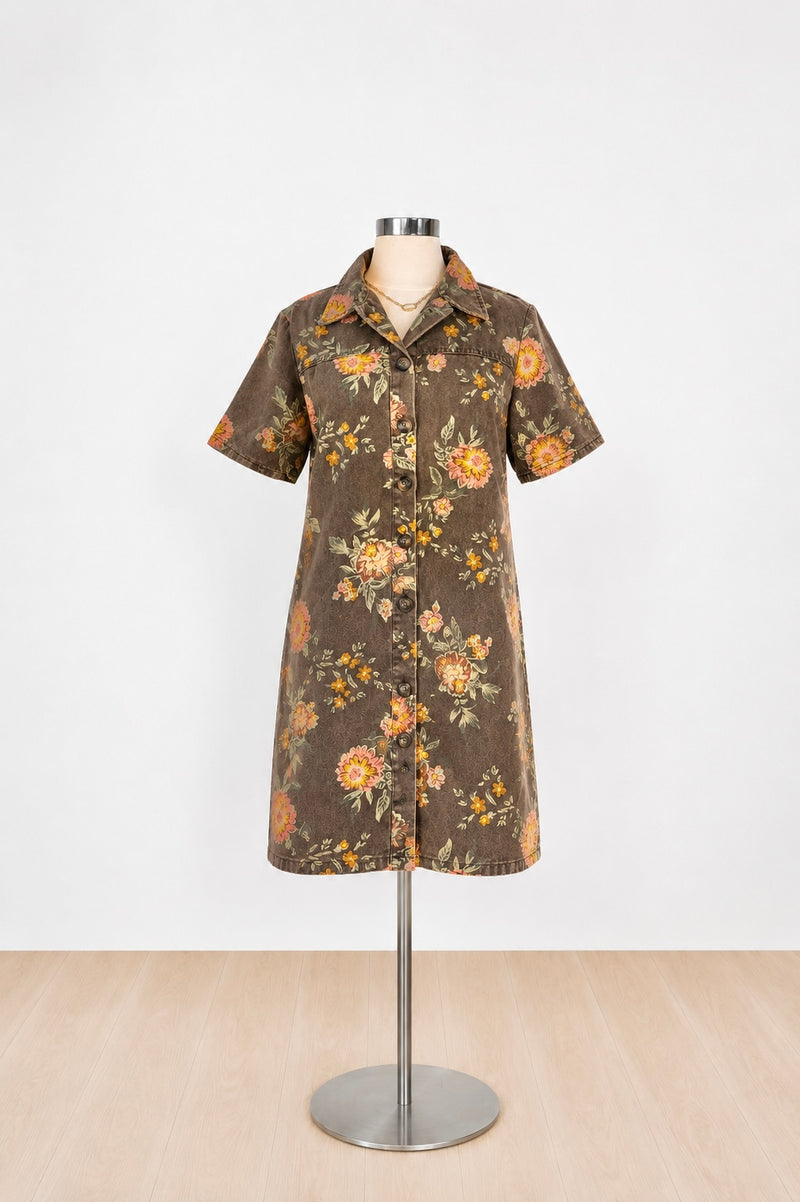 A short-sleeve button-front shirt dress displayed on a mannequin stand. The dress has a brown base with a retro floral pattern in orange, yellow, and green, set against a plain white wall and light wood floor.