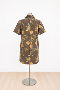A short-sleeve button-front shirt dress displayed on a mannequin stand, featuring a brown base with a yellow and orange floral pattern and green leaves, set against a white wall and light wood floor.