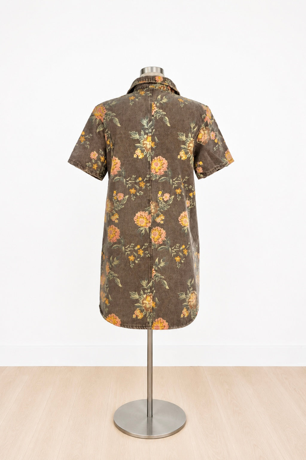 A short-sleeve button-front shirt dress displayed on a mannequin stand, featuring a brown base with a yellow and orange floral pattern and green leaves, set against a white wall and light wood floor.