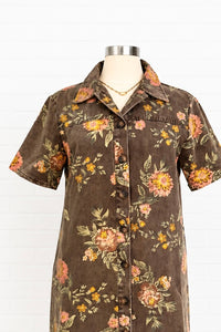 A short-sleeve button-front shirt dress displayed on a mannequin torso against a white paneled background. The dress has a dark brown base with a vintage floral print in pink, orange, yellow, and green.