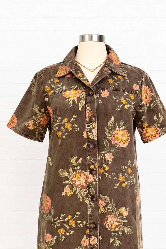 A short-sleeve button-front shirt dress displayed on a mannequin torso against a white paneled background. The dress has a dark brown base with a vintage floral print in pink, orange, yellow, and green.