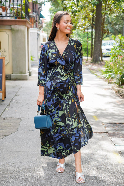 Model is wearing a black, blue, and green maxi dress with siver heels and a blue purse on a downtown street.