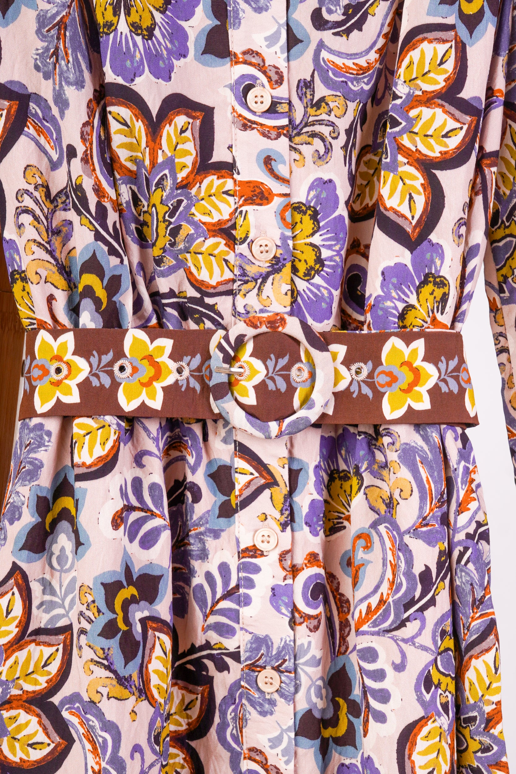 A close-up of a patterned dress or garment with a wide brown belt featuring a matching floral print. The fabric has an abstract, painterly pattern in purple, orange, yellow, and dark brown on a light background.