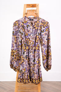 A long-sleeve button-front dress with a ruffled waist and a paisley floral print, hanging on a wooden hanger on a wooden chair against a white wall.