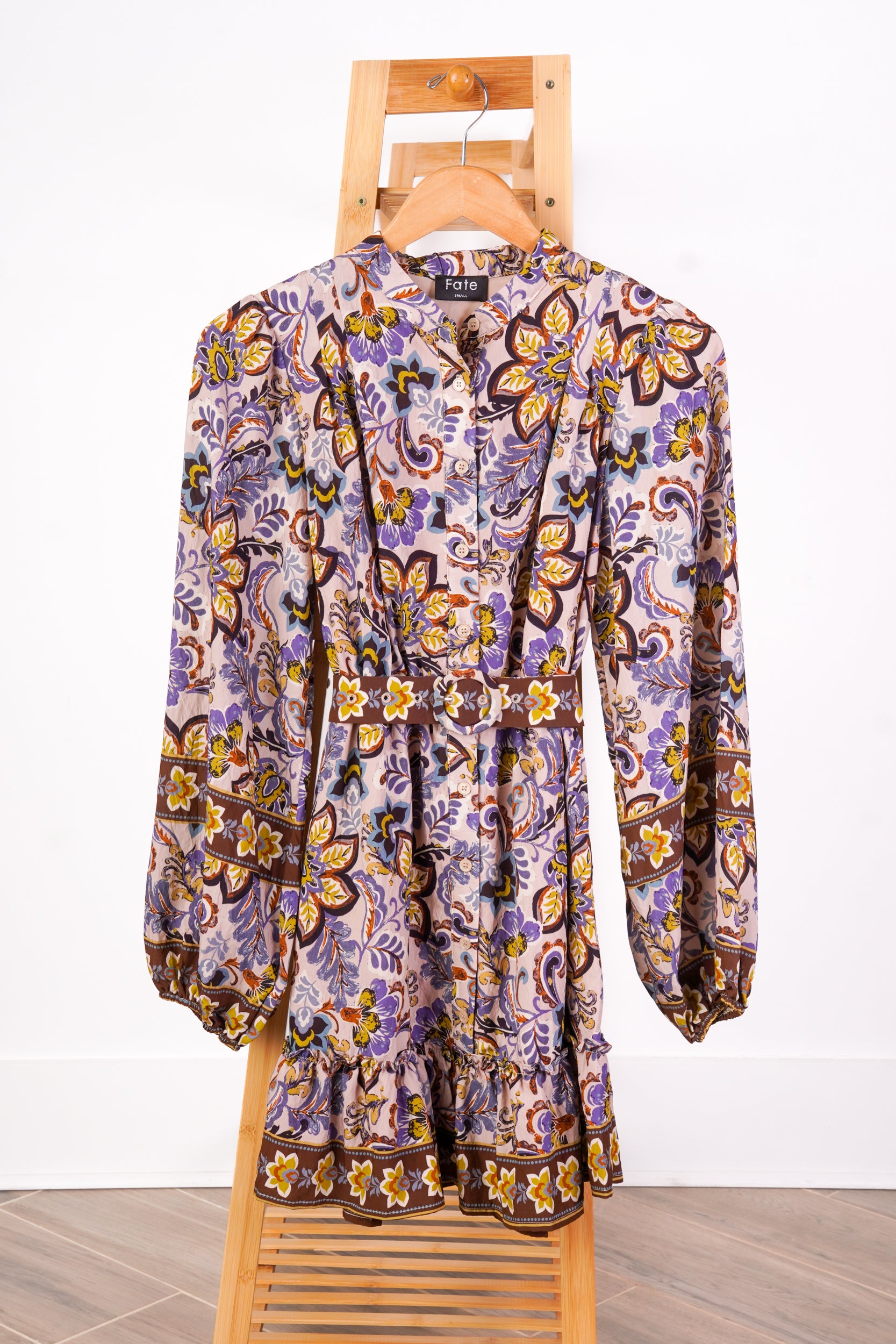 A long-sleeve button-front dress with a ruffled waist and a paisley floral print, hanging on a wooden hanger on a wooden chair against a white wall.