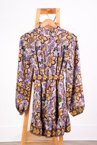 A long-sleeve bohemian-style mini dress with an intricate paisley and floral pattern in purple, yellow, brown, and white, hanging on a wooden hanger from a wooden display stand against a white wall.