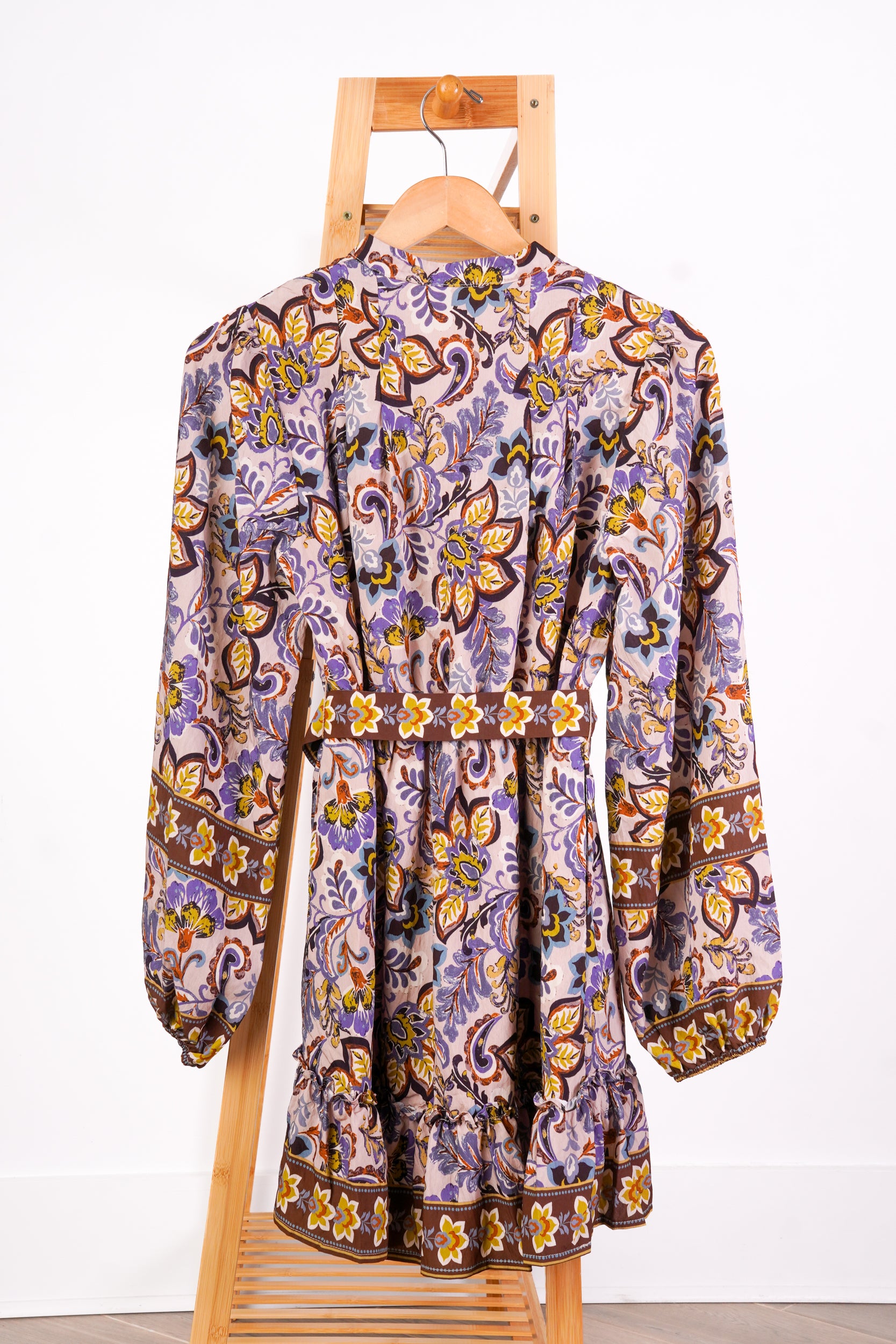 A long-sleeve bohemian-style mini dress with an intricate paisley and floral pattern in purple, yellow, brown, and white, hanging on a wooden hanger from a wooden display stand against a white wall.