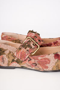 A close-up of a pair of women's flat shoes featuring a woven floral pattern in muted red, green, and beige tones, accented with a wide strap and a gold-tone buckle.
