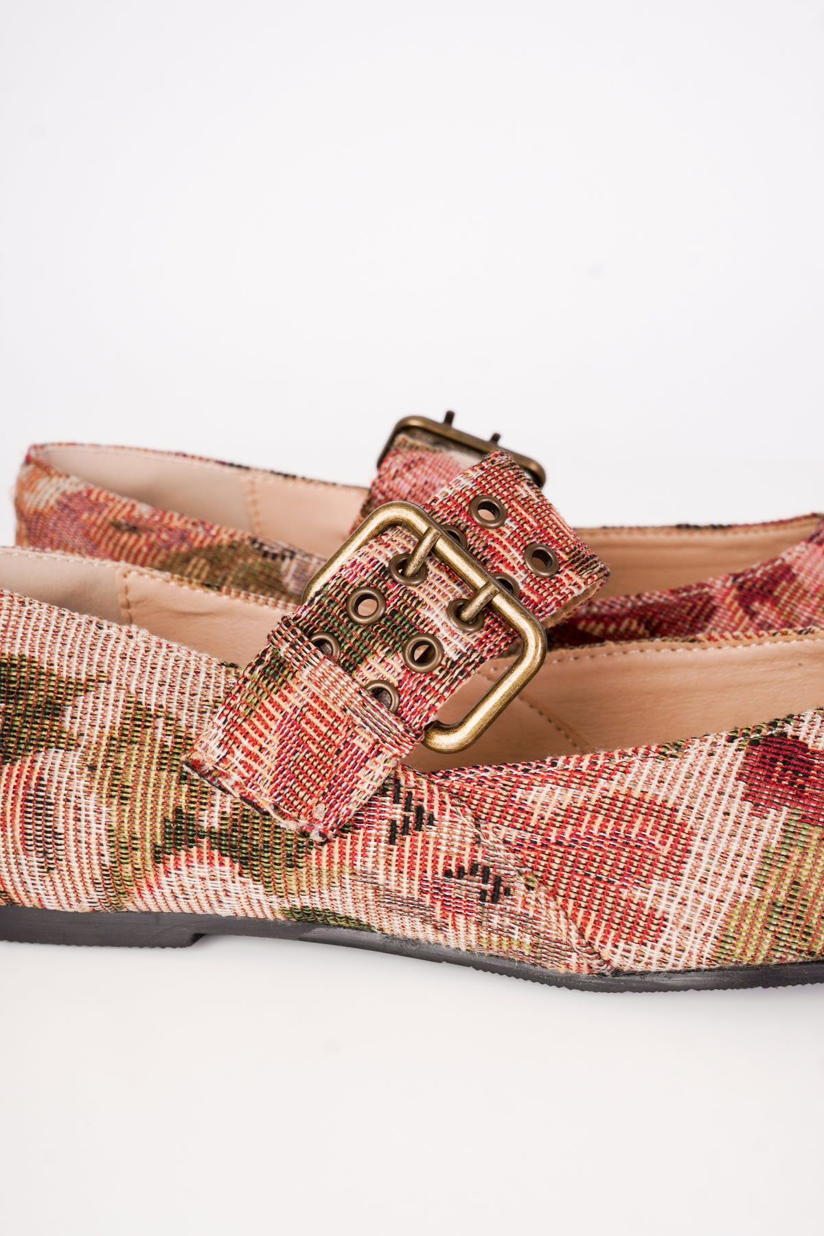 A close-up of a pair of women's flat shoes featuring a woven floral pattern in muted red, green, and beige tones, accented with a wide strap and a gold-tone buckle.