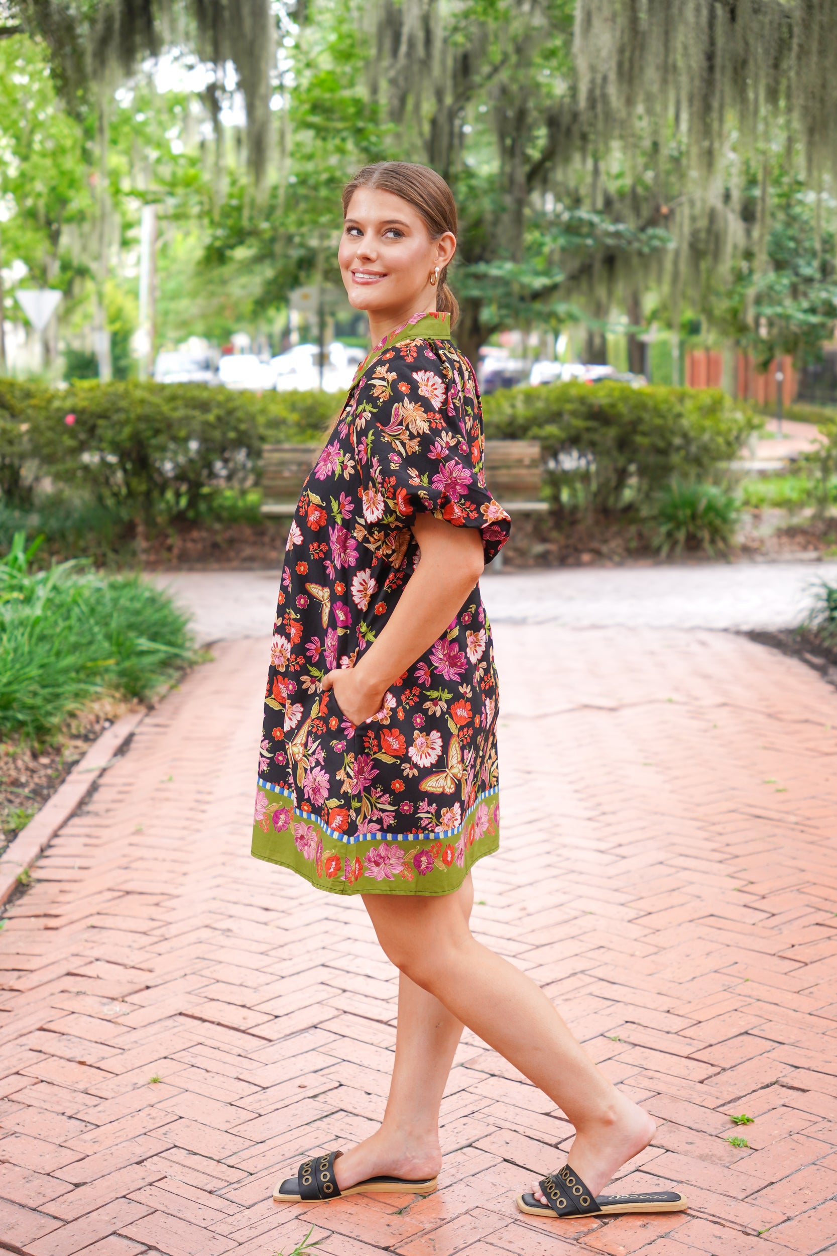 A woman wearing a short-sleeve black floral dress with a green border walks along a brick pathway in a park with trees and hanging moss in the background.