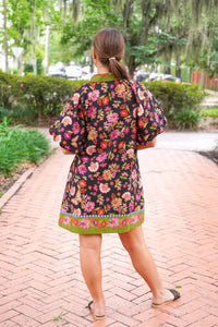 A woman viewed from behind wearing a short puff-sleeve dress with a colorful floral print on a black background, standing on a brick pathway in a park.