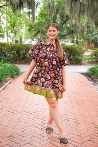A woman wearing a black floral short-sleeve dress with a green collar and hem, walking on a brick pathway in a park with Spanish moss trees in the background.