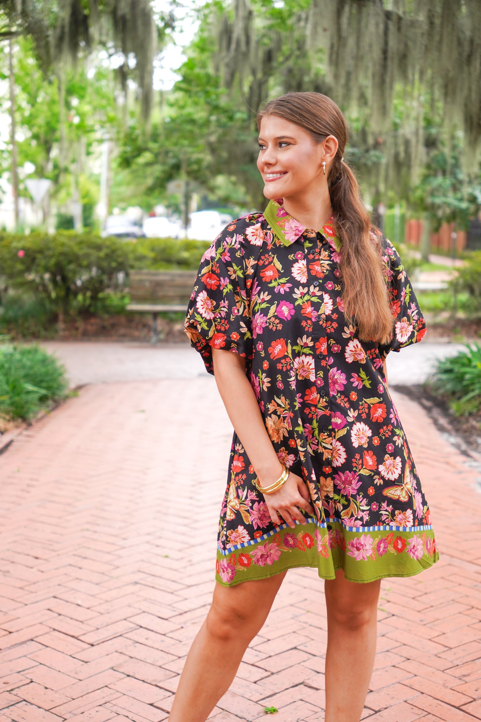 A woman wearing a short-sleeve black floral dress with a green collar and hem, standing on a brick pathway in a park with trees and hanging moss in the background.