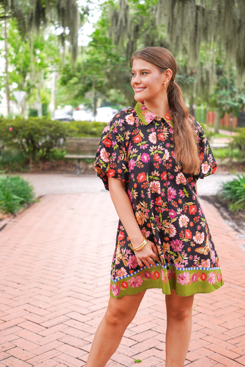 A woman wearing a short-sleeve black floral dress with a green collar and hem, standing on a brick pathway in a park with trees and hanging moss in the background.