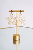 Photo shows a pair of drop earrings with white flowers on a gold earring stand