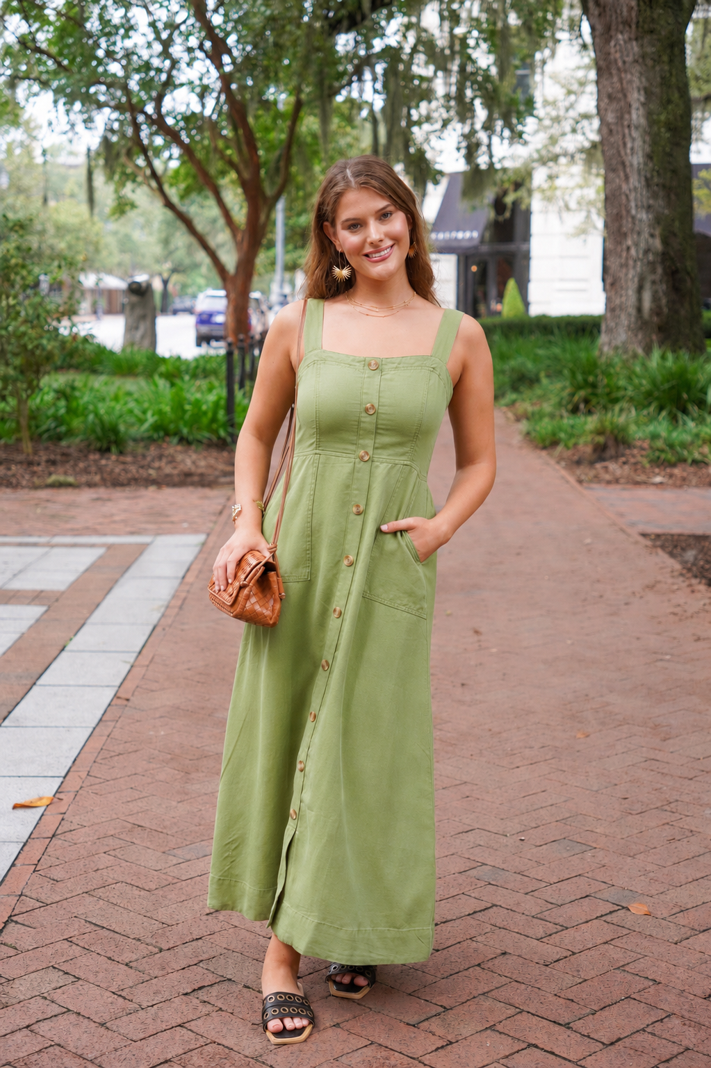 A woman wearing a sage green linen maxi dress with a button-down front and side pockets, standing on a brick pathway in a park setting.