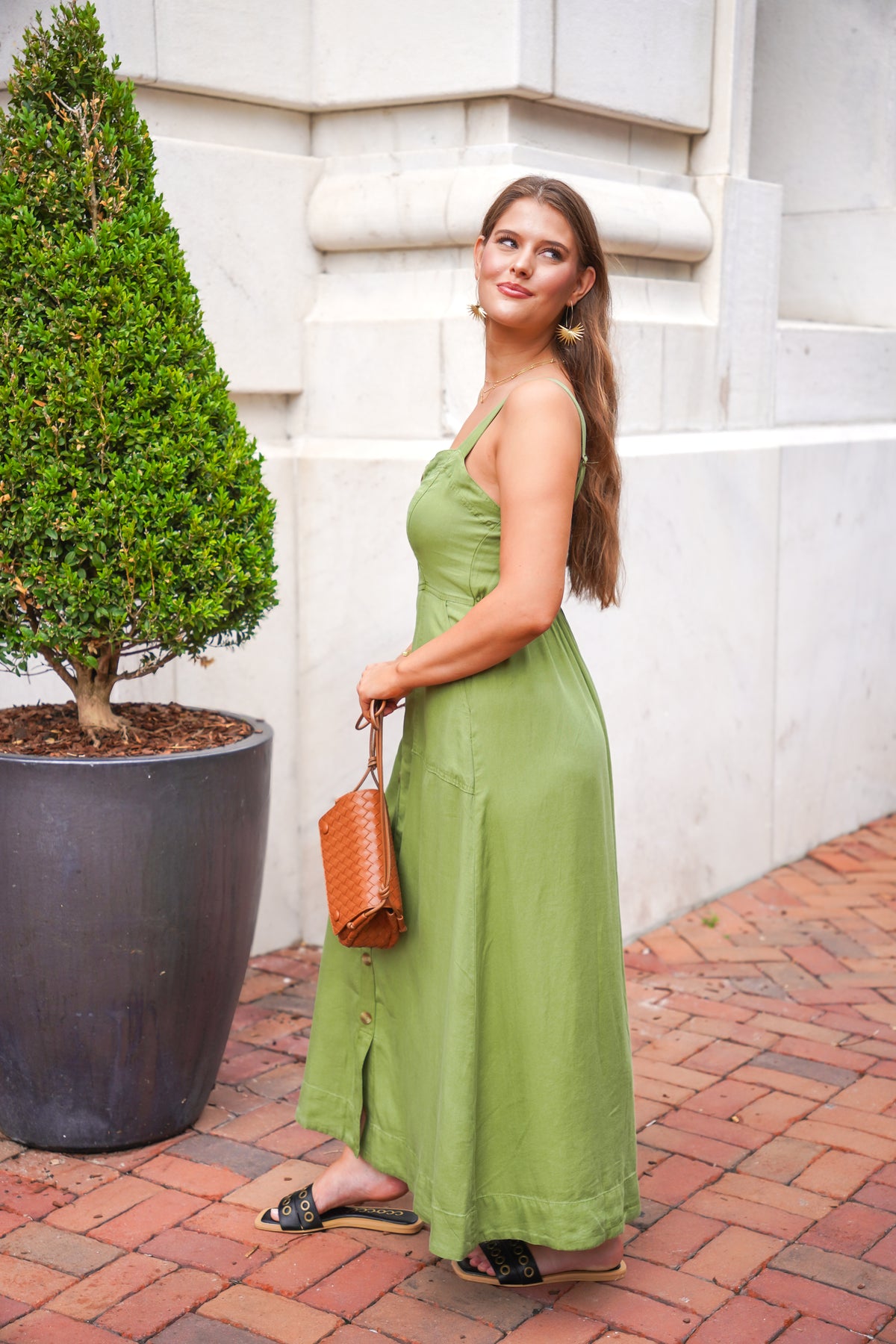 A woman wearing a sage green maxi dress stands on a brick sidewalk next to a large potted tree, holding a brown woven handbag.