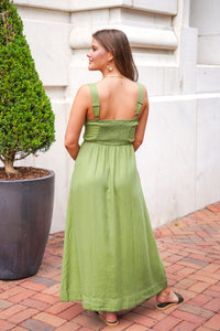 A woman wearing a light olive green linen maxi dress with a smocked bodice and adjustable shoulder straps, photographed from behind on a brick walkway next to a potted tree.