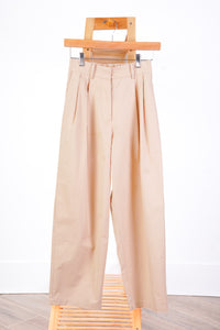 A pair of high-waisted, wide-leg pleated trousers in a light beige or tan color, hanging from a wooden hanger on a white wall.