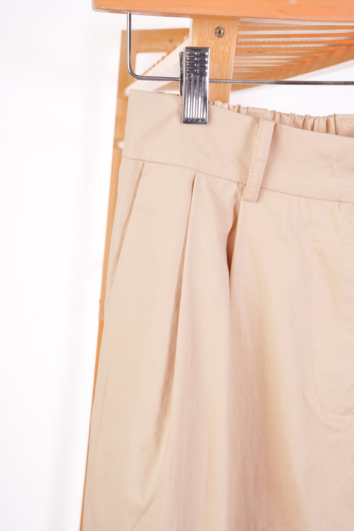 Close-up of beige pleated trousers hanging from a metal hanger with a wooden rack behind them.