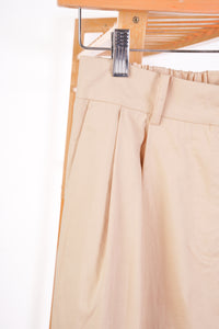 Close-up of beige pleated trousers hanging from a metal hanger with a wooden rack behind them.