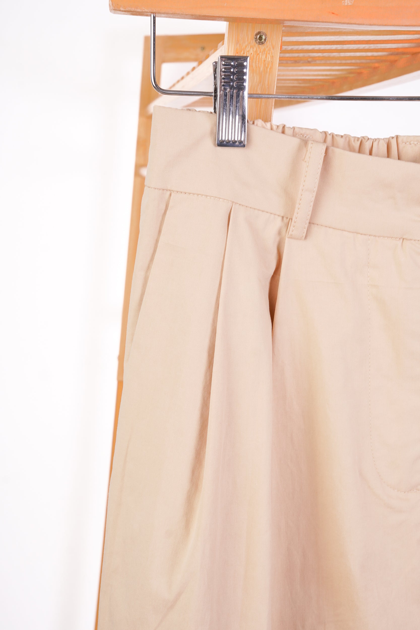 Close-up of beige pleated trousers hanging from a metal hanger with a wooden rack behind them.