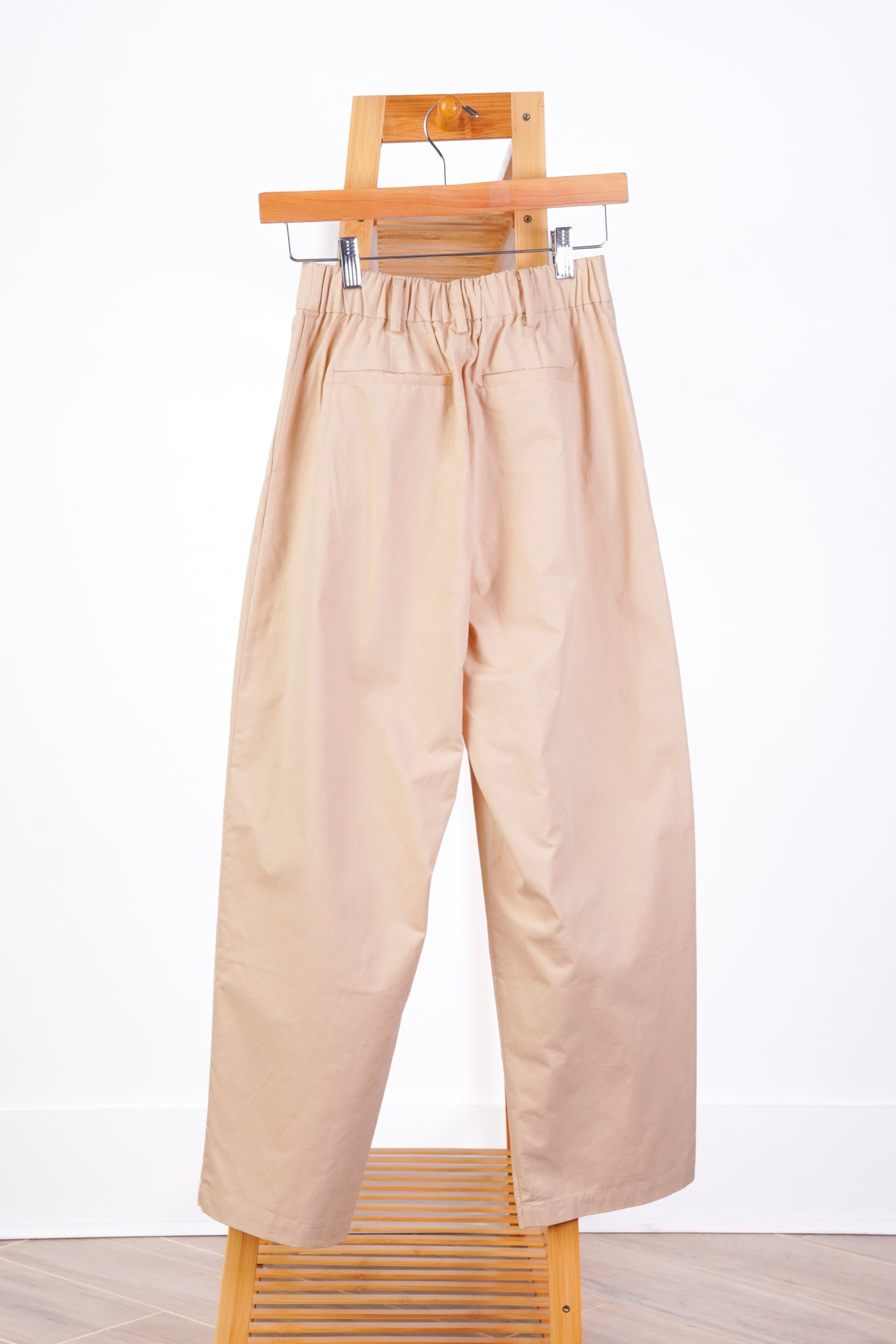 A pair of wide-leg, cropped beige trousers with an elastic waistband, hanging from a wooden hanger on a wooden stand against a white wall.