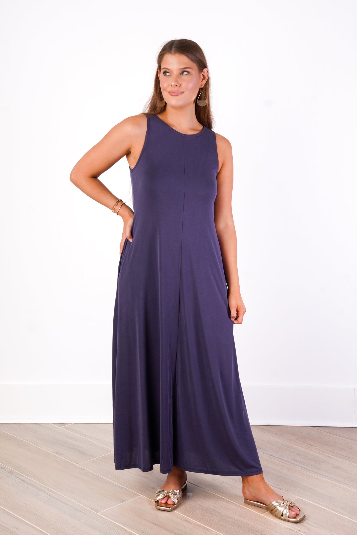 A woman modeling a sleeveless, floor-length navy maxi dress with side pockets, paired with gold flat sandals, standing against a white wall and light wood floor.