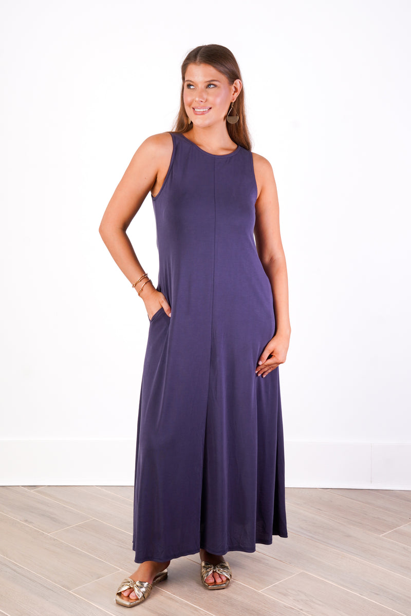 A woman modeling a sleeveless, floor-length purple maxi dress with side pockets, standing against a white wall with light wood flooring.