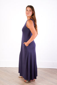 A woman modeling a sleeveless, floor-length navy maxi dress with side pockets, standing in profile against a white wall and light wood floor.