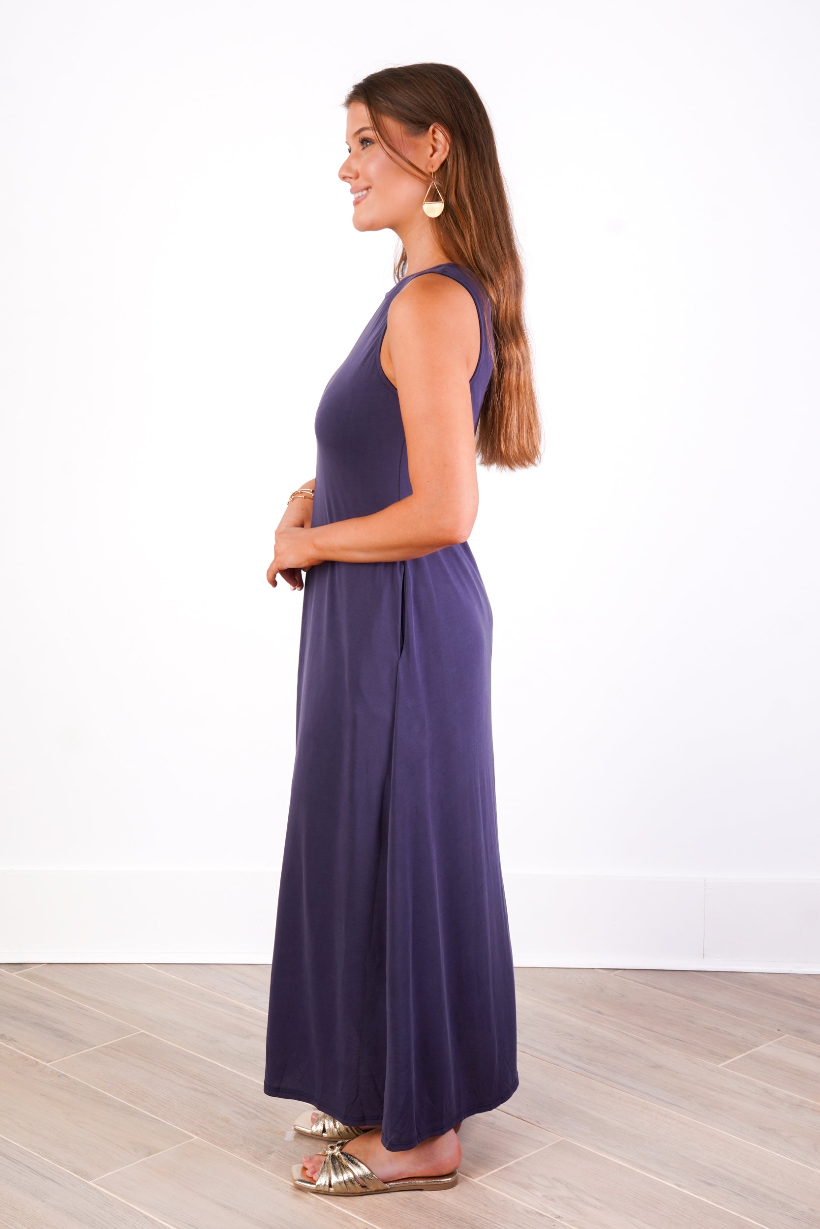 A woman modeling a sleeveless, floor-length navy maxi dress, shown in profile against a white wall and light wood floor.