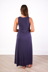 A woman viewed from behind wearing a sleeveless, floor-length dark purple maxi dress, standing on a light wood floor against a white wall.