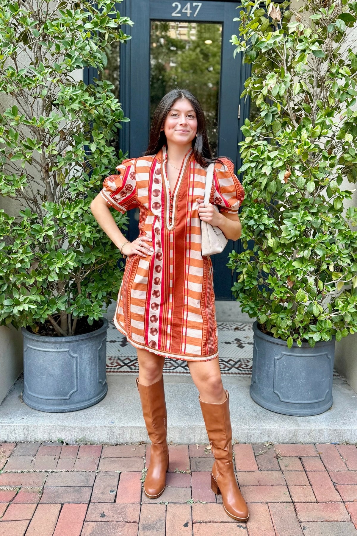 A woman stands outdoors in front of a dark door framed by leafy green plants, wearing a short puff-sleeve dress with orange and white vertical stripes and red circular motifs, paired with brown knee-high boots and a light beige shoulder bag.